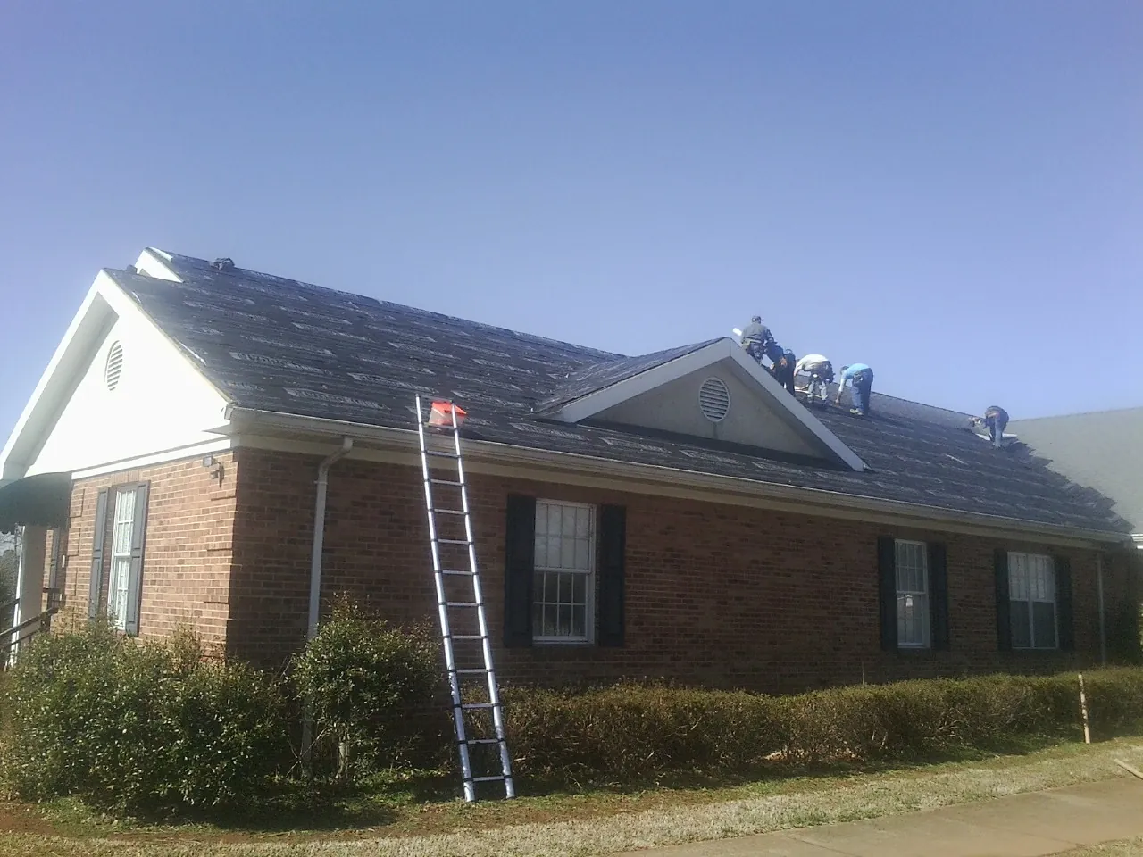 Crew tearing shingles off a brick ranch roof with a ladder set — Emergency Roof Repair work by our crew in Olyphant