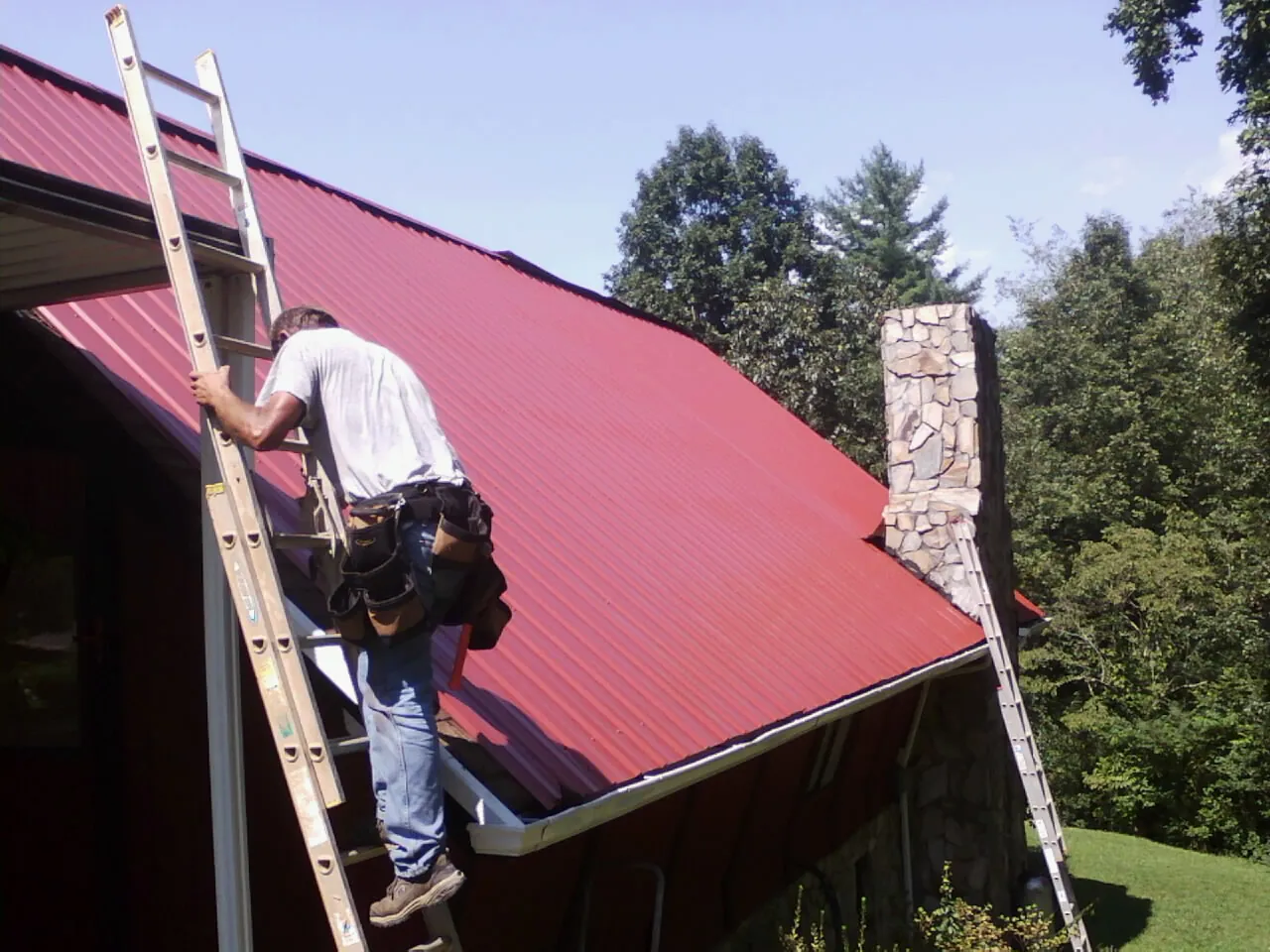 Roofer on a ladder beside a red metal roof and stone chimney — Soffit & Fascia Repair by Roofing Brothers in Olyphant
