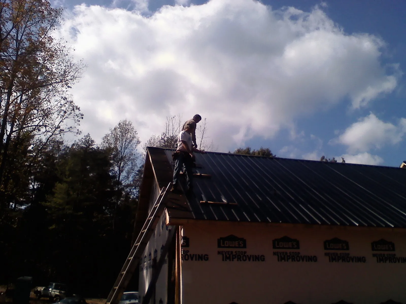 Roofer fastening dark metal panels high against a cloudy sky, from a Roof Inspection job in Olyphant